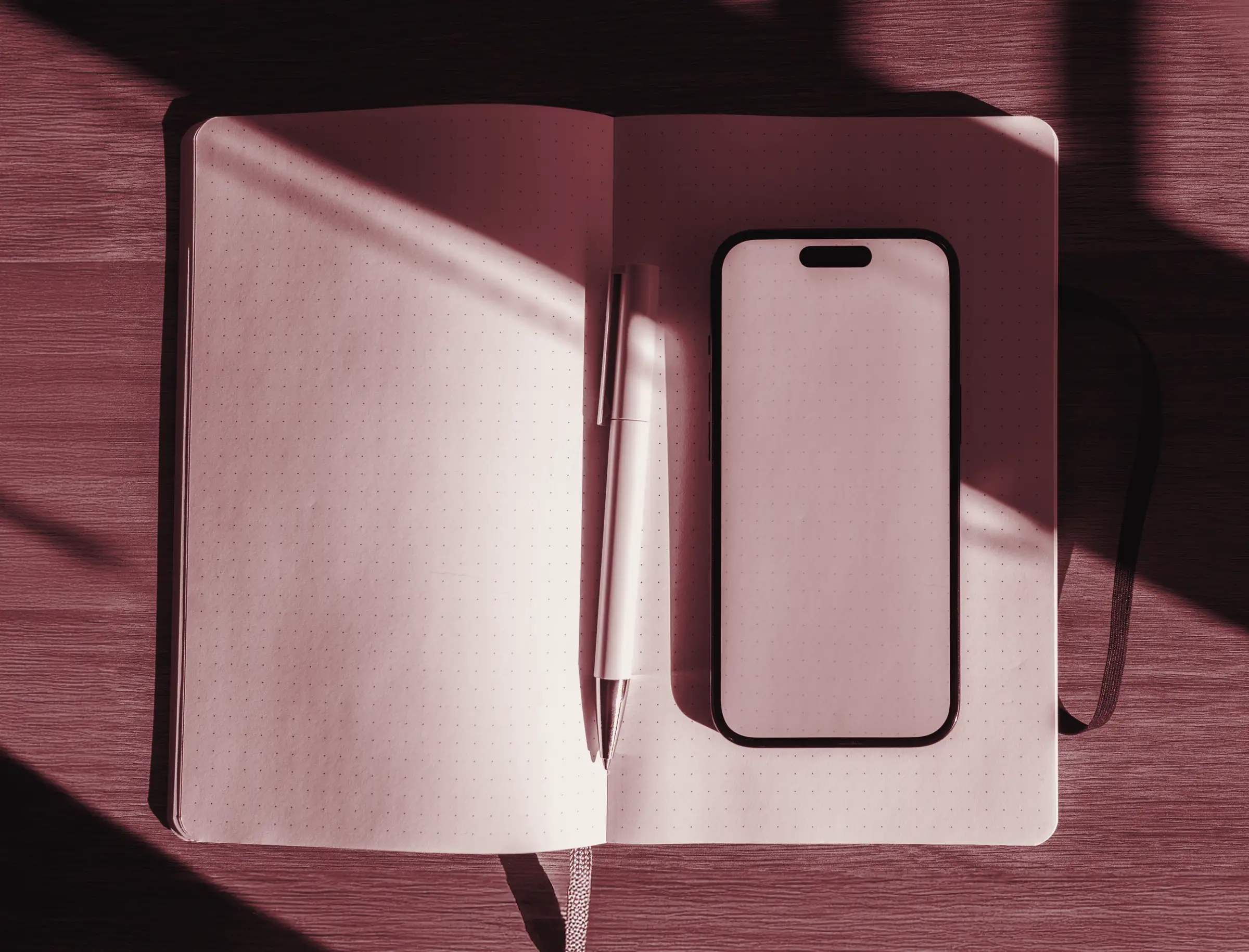 Quaderno a quadretti aperto con una penna e uno smartphone dallo schermo vuoto, su un tavolo di legno attraversato da strisce di sole