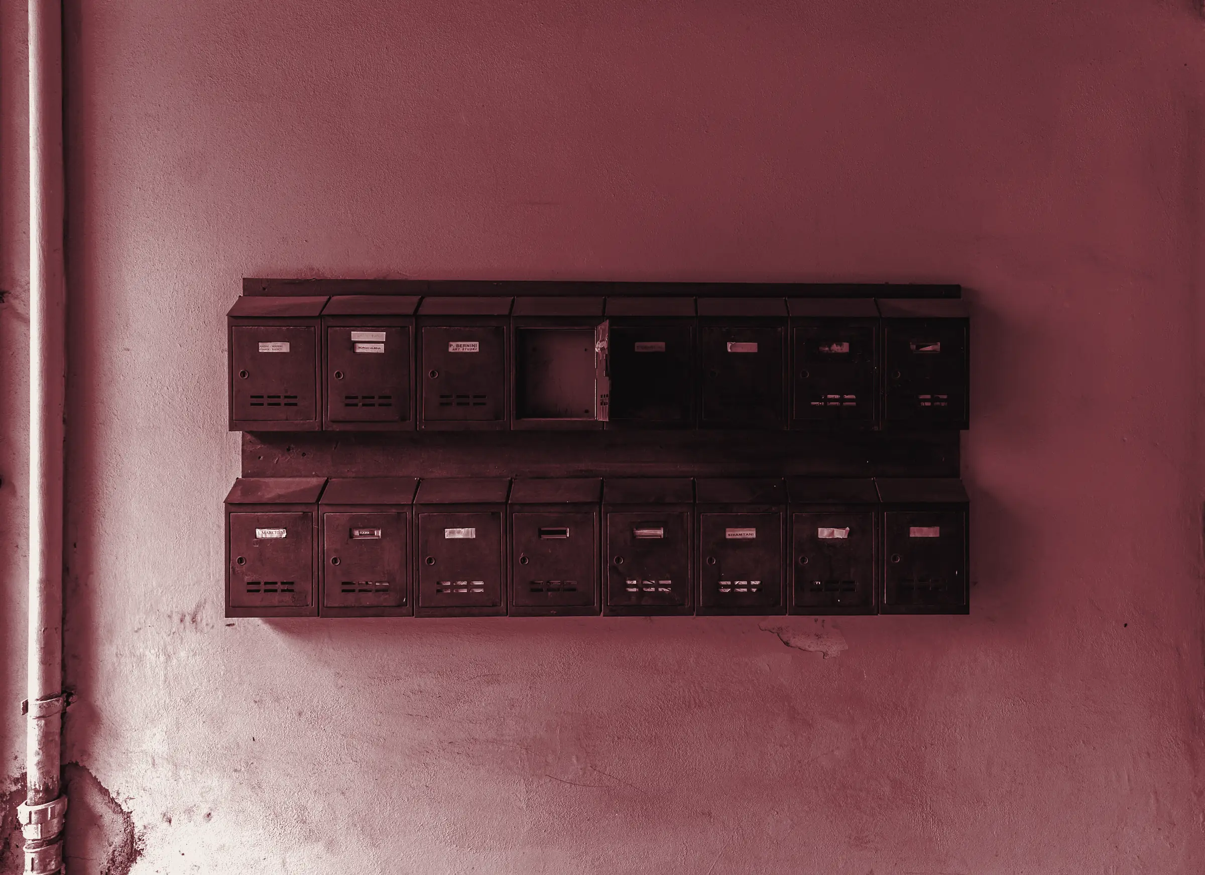 Fila di cassette postali scure appese a una parete color ocra, con uno sportello aperto e vuoto