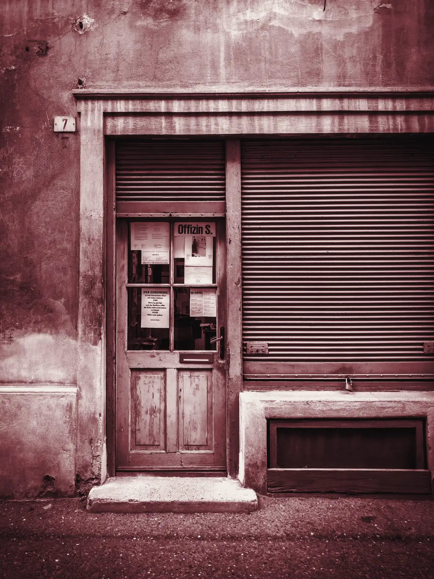 Porta di legno con vetri e saracinesca abbassata di una bottega su un muro color ruggine, in una strada italiana