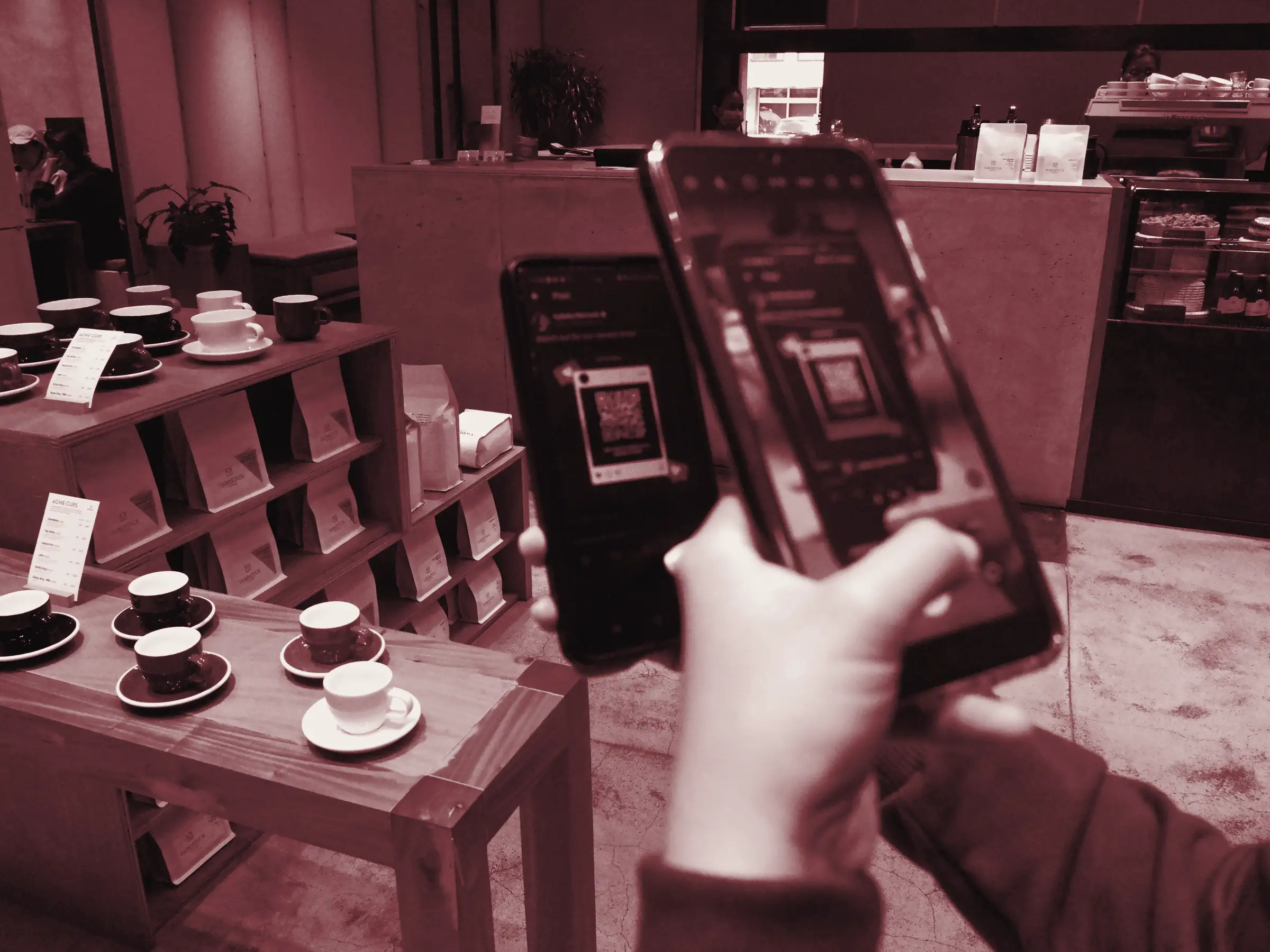Due mani tengono due smartphone davanti al bancone di un caffè, uno inquadra un codice QR, sullo sfondo scaffali di legno con tazzine